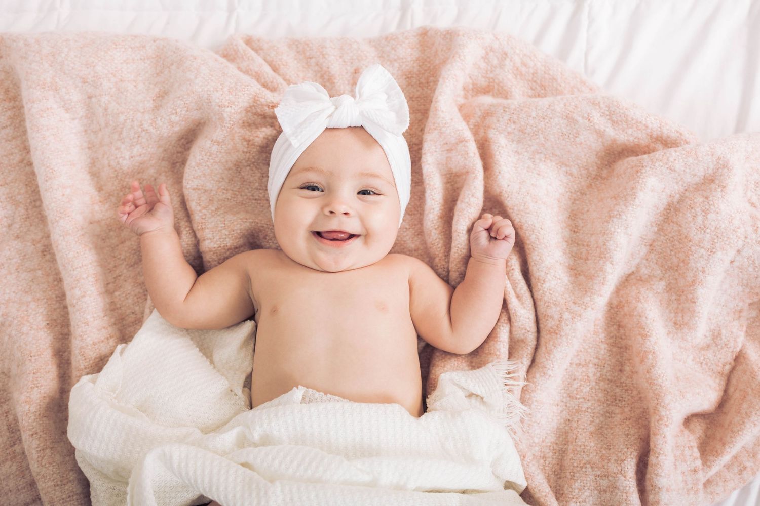 A smiling baby wearing a white bow headband lying on pink blankets.