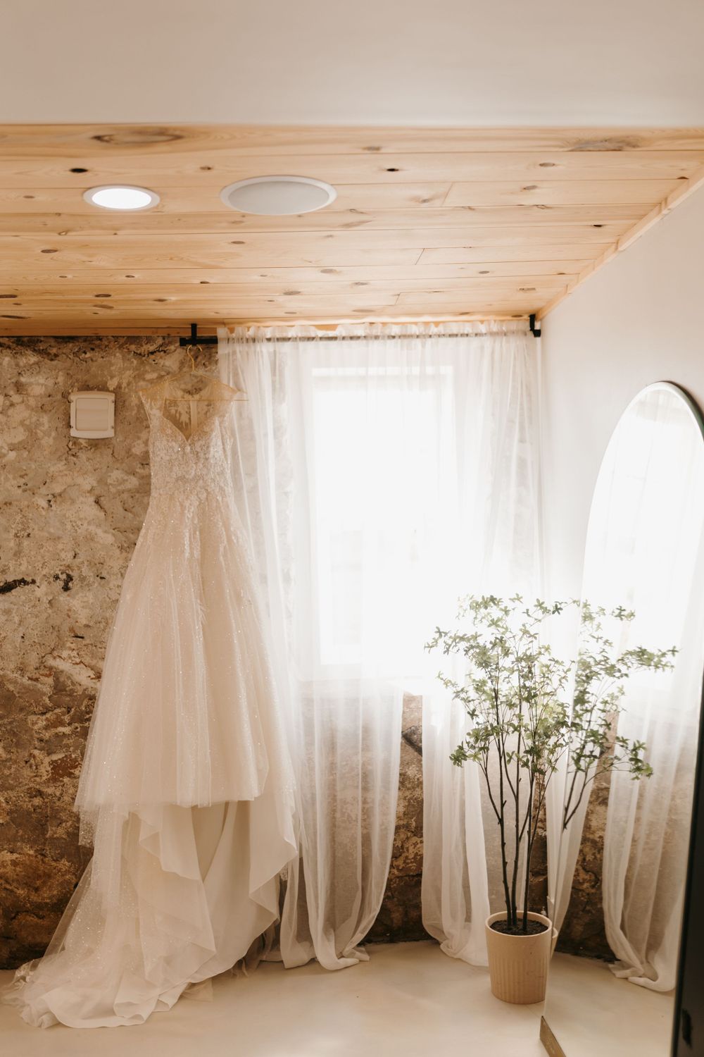 Elegant wedding dress hanging by a sunlit window in a rustic bridal suite with wooden ceiling.