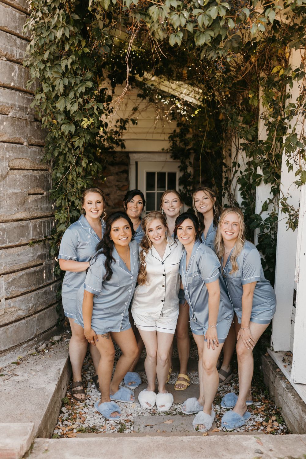 Bride in white pajamas poses with eight bridesmaids in matching blue rompers outdoors.