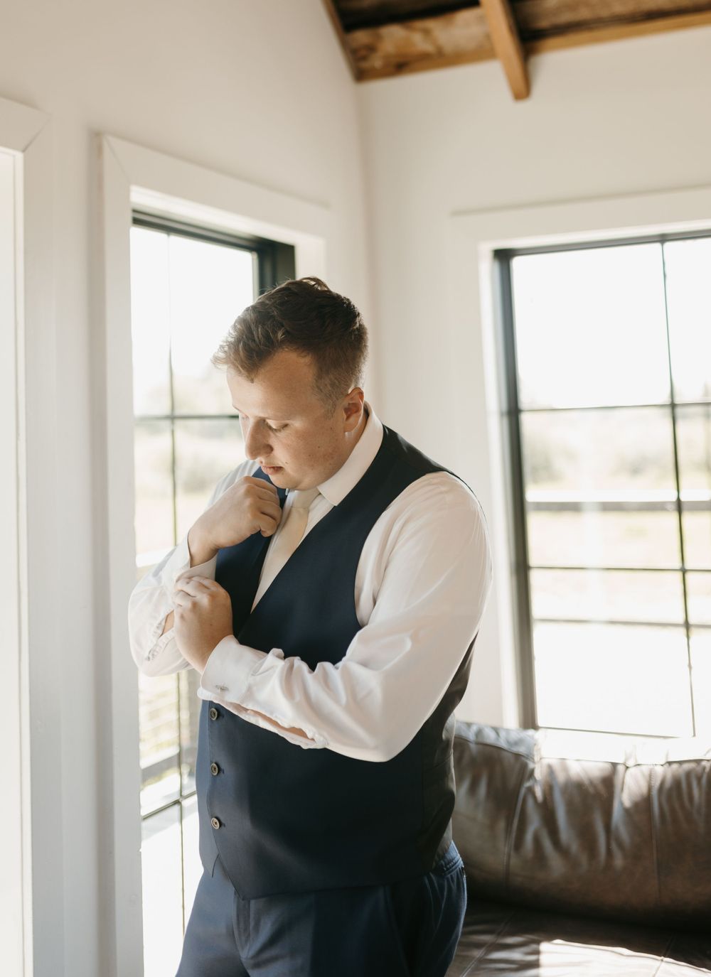 Groom in navy vest and white shirt adjusting his cufflinks by a sunlit window.