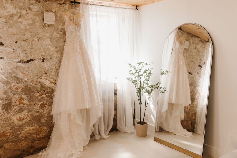 Bridal gown displayed near a large arched mirror and potted plant in a rustic stone-walled room.