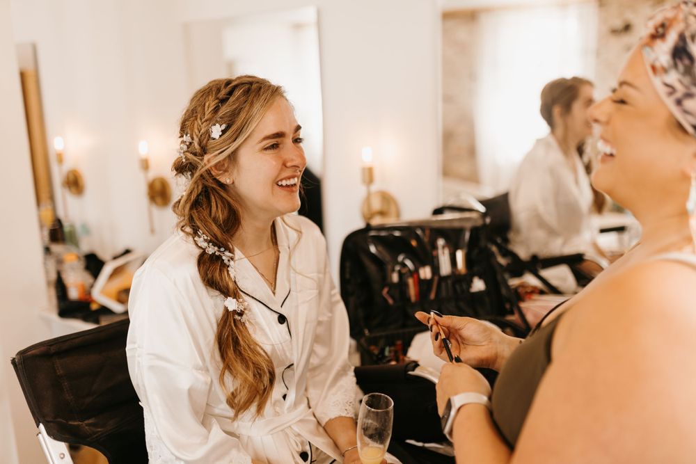 Bride laughing joyfully with her makeup artist during wedding morning preparations in a bright suite.
