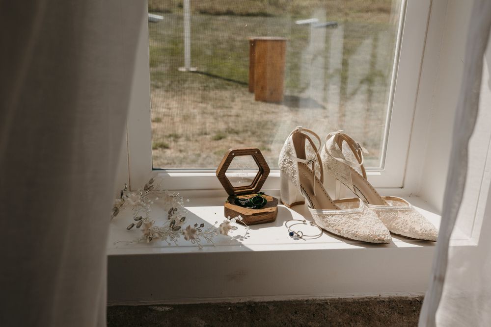 Lace bridal heels and hexagonal wooden ring box displayed on a sunlit white window sill.
