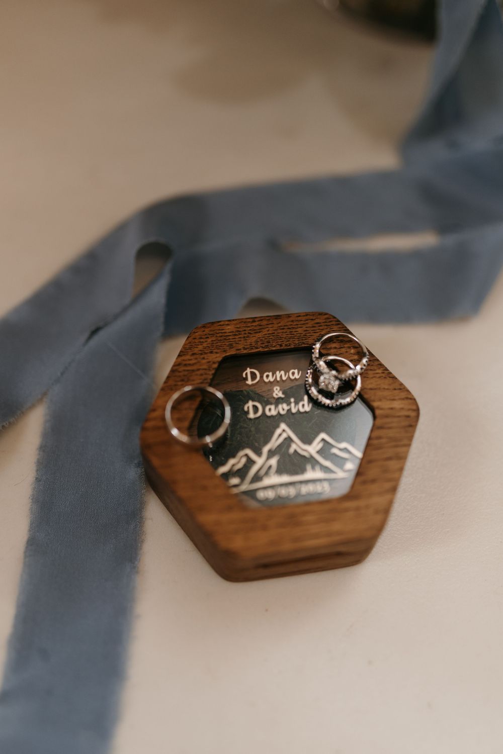Personalized hexagonal wooden ring box engraved with Dana and David beside wedding rings and blue ribbon.