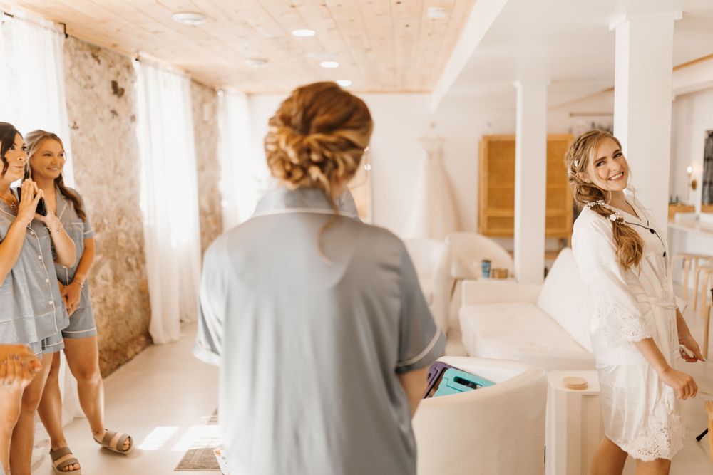 Bride in white robe smiling at bridesmaids getting ready in bright bridal suite.