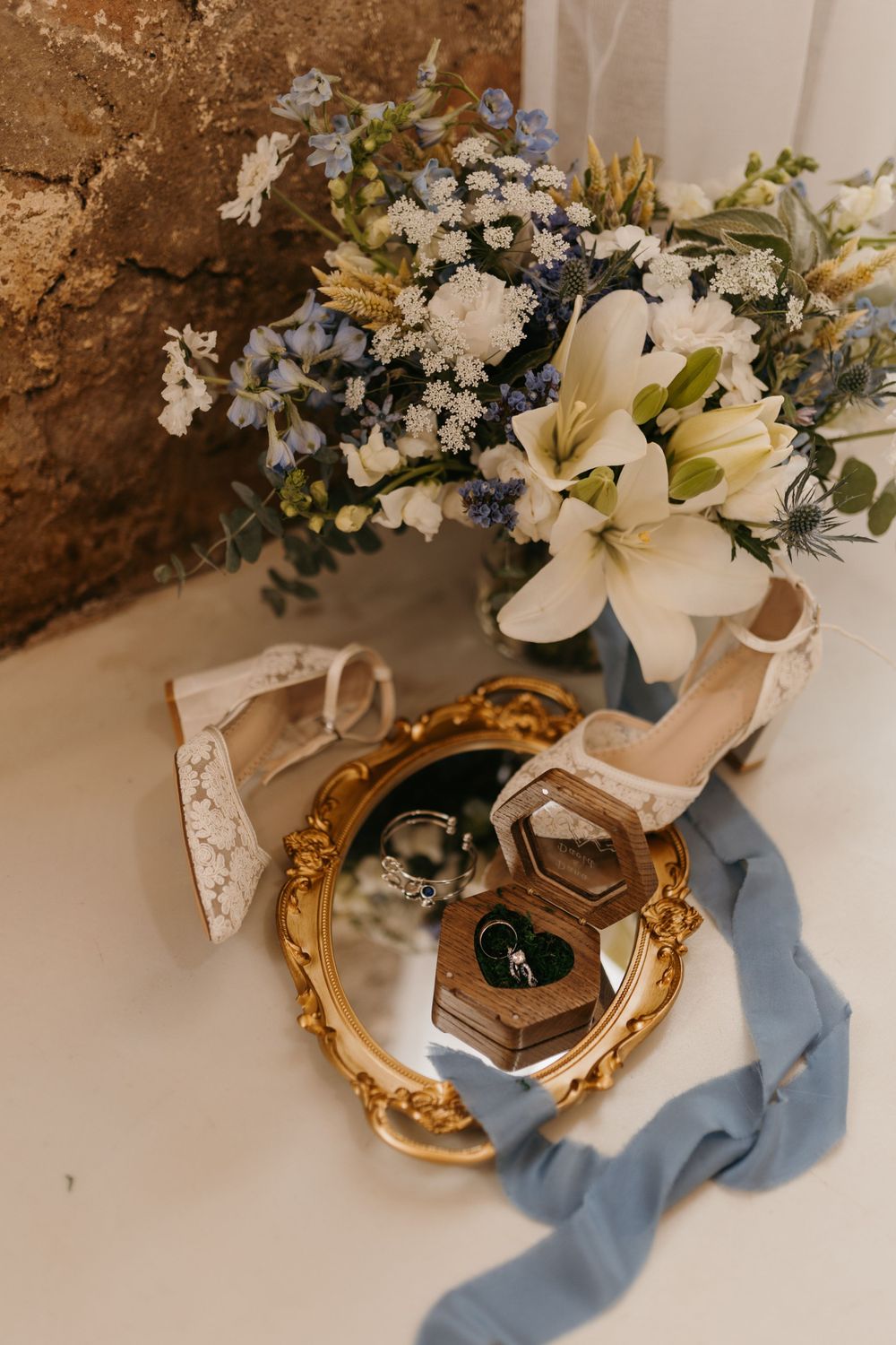 Wedding flat lay with white and blue floral bouquet, lace heels, gold mirror, and ring box.