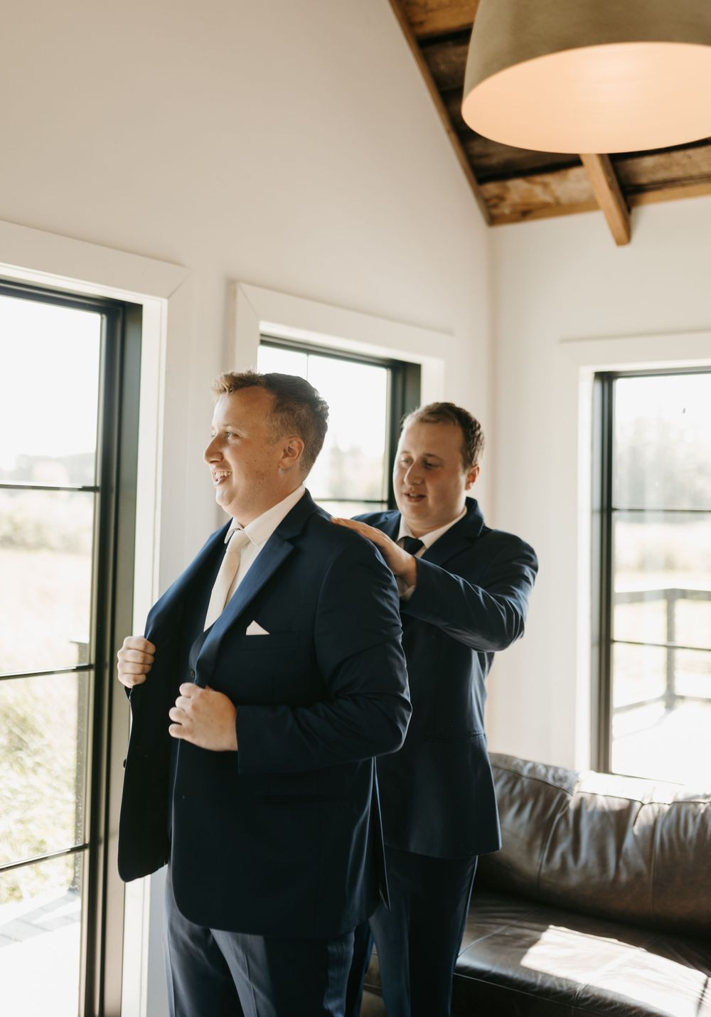 Groomsman helping the groom put on his navy wedding suit jacket in a bright venue.