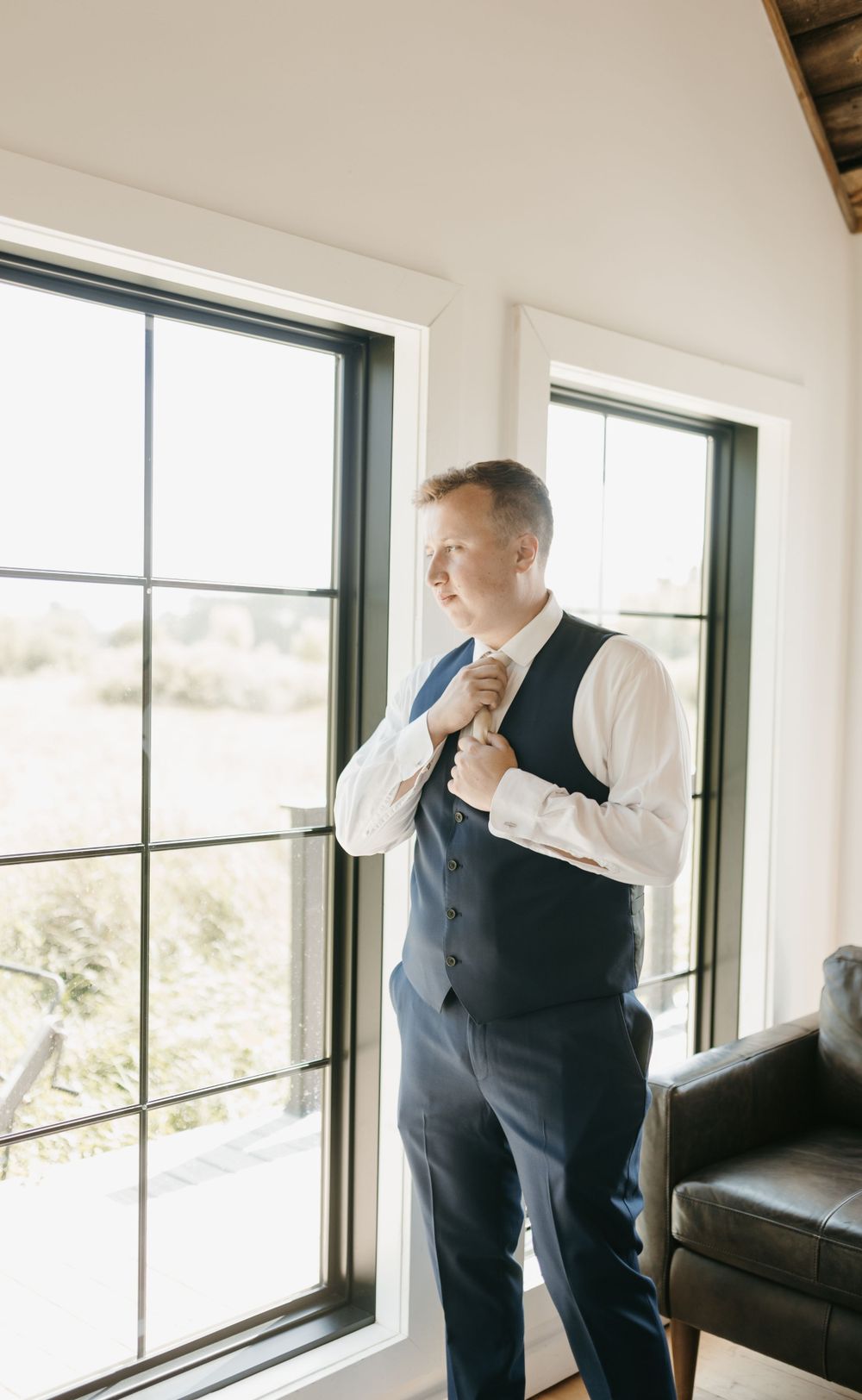 Dapper groom adjusting his tie while standing by large windows in a bright modern room.