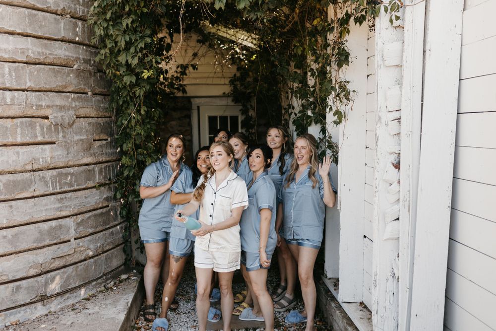 Bride and bridesmaids in matching blue rompers laughing together in ivy-covered doorway.