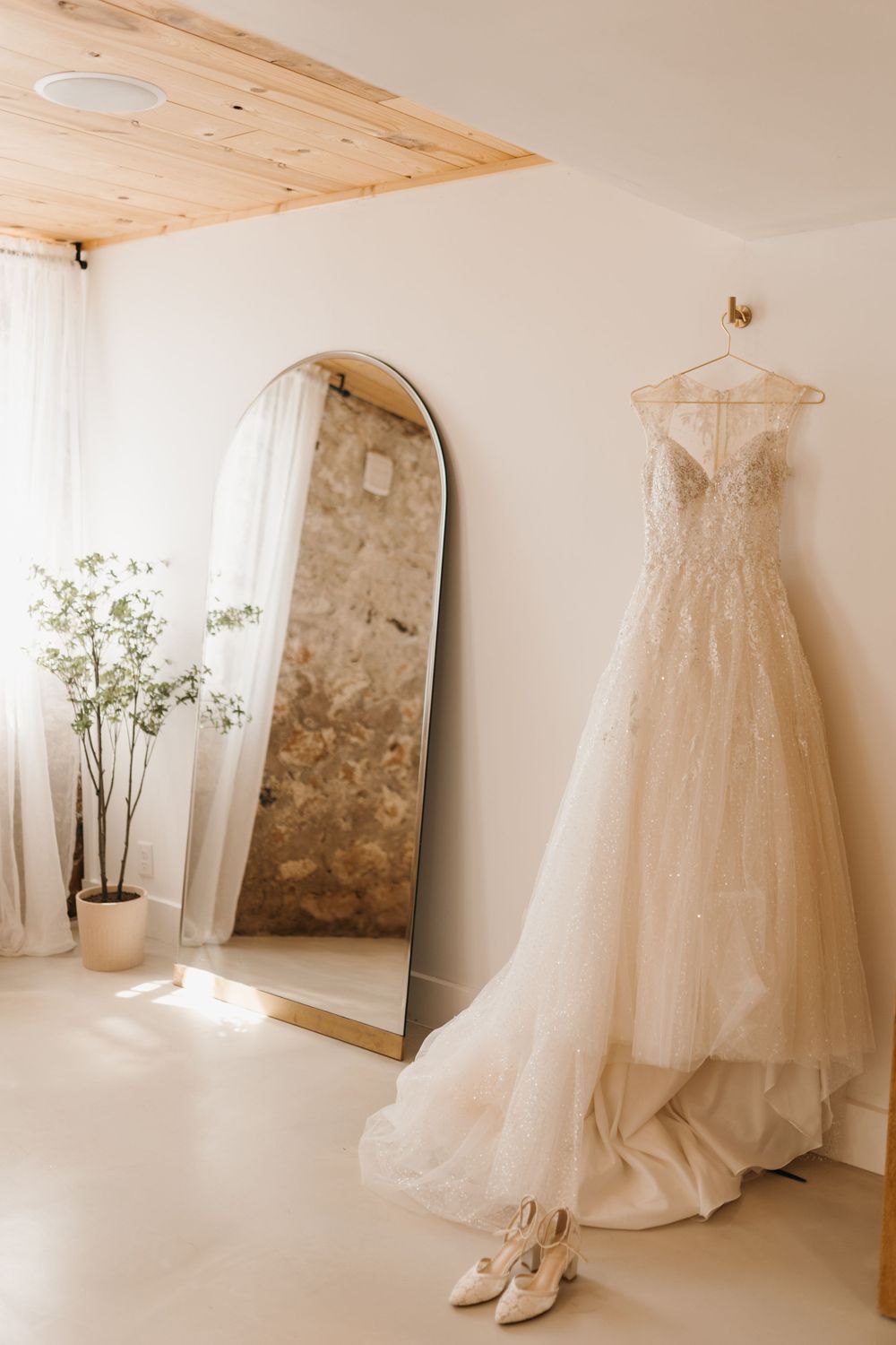 Lace wedding gown hanging on a wall beside a large arched floor mirror in a bright bridal suite.