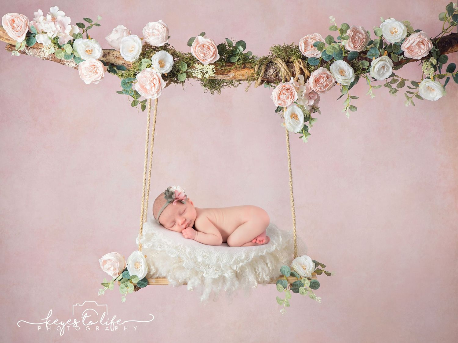 Sleeping newborn on floral swing with pink and white roses against pink background.