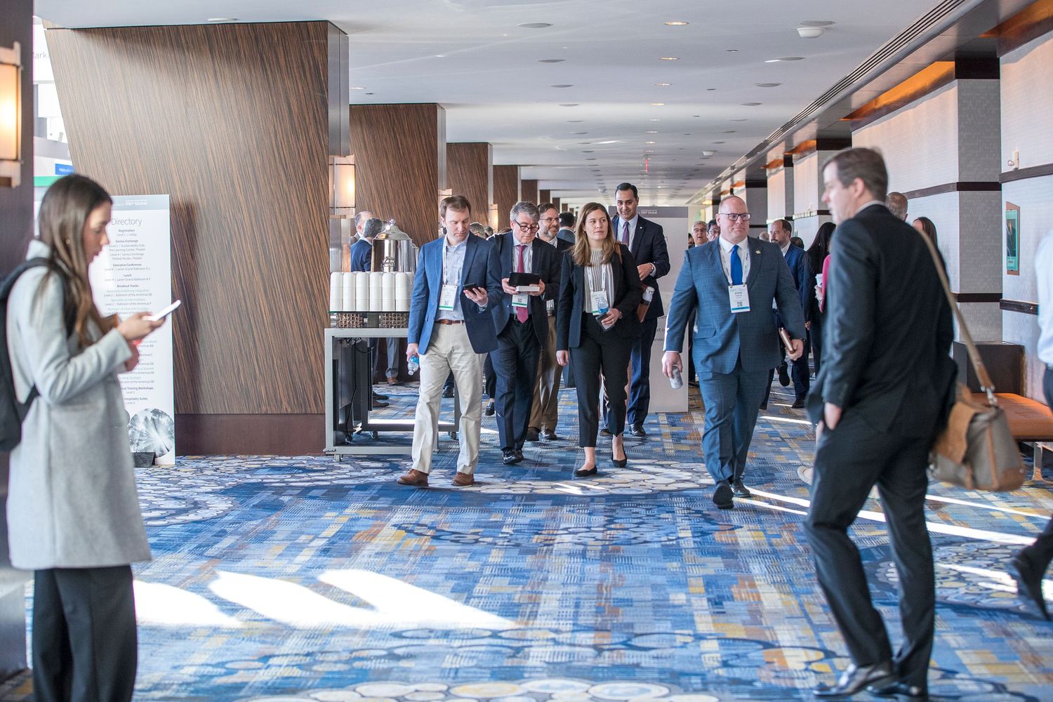 Business professionals networking in a large hallway with blue carpeting at a corporate event.