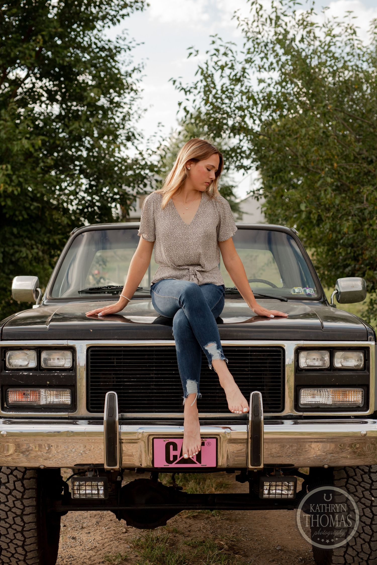 Person in casual attire poses on the hood of a vintage black truck against a leafy green background.