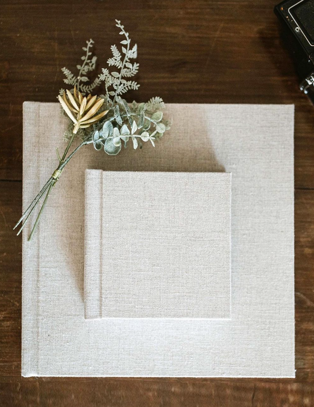 White linen photo album with dried eucalyptus arrangement on wooden surface.