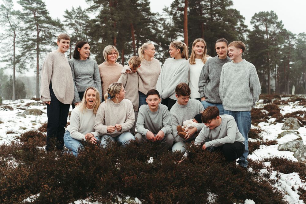 A casual group photo of people in matching neutral sweaters posing together in a winter forest setting.