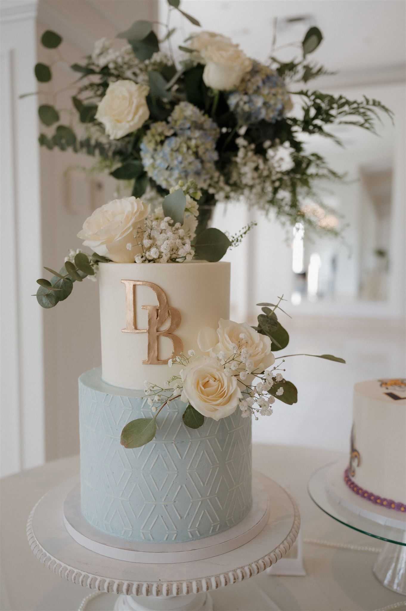 Two-tier wedding cake with white and blue geometric design decorated with fresh roses and eucalyptus.