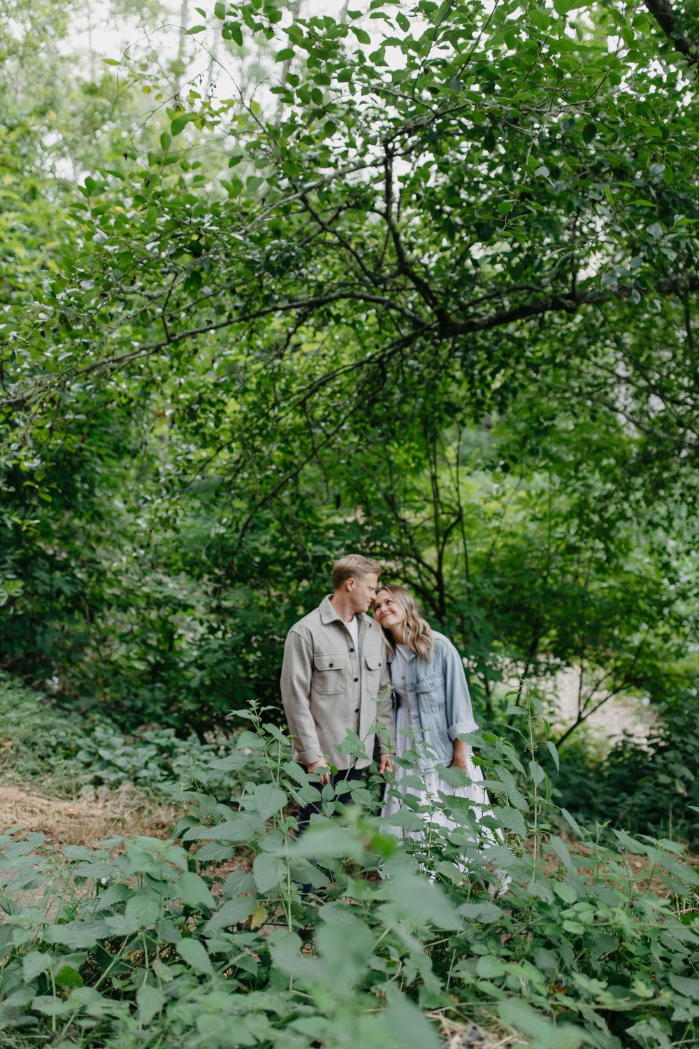 A couple shares an intimate moment together under lush green trees in a forest setting.