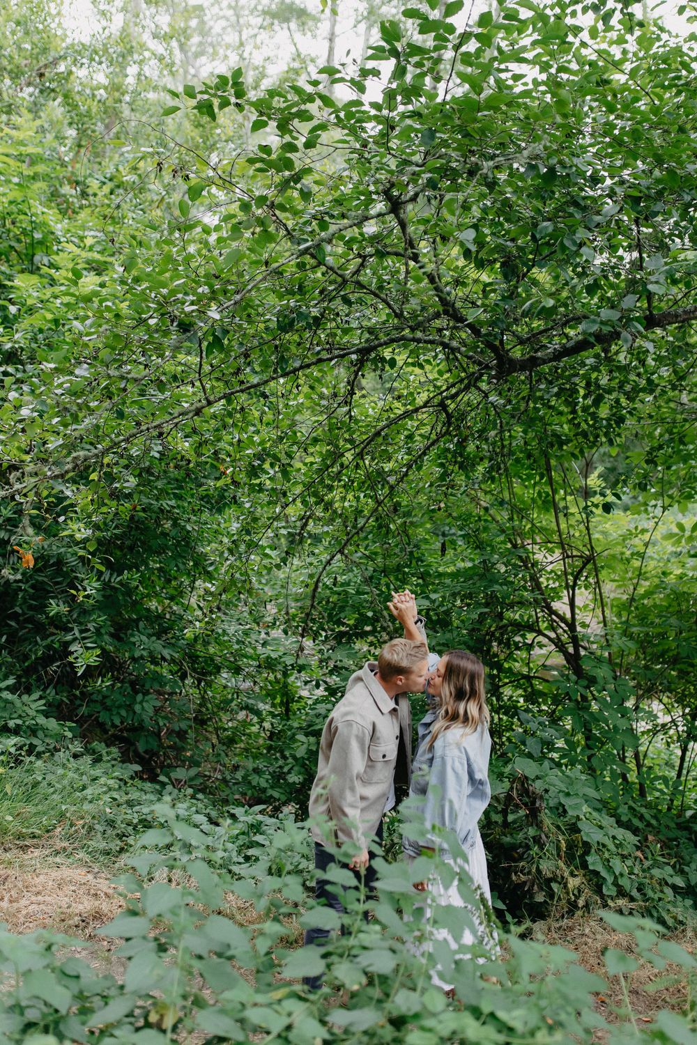 A romantic moment captured between a couple beneath overhanging tree branches in a forest.