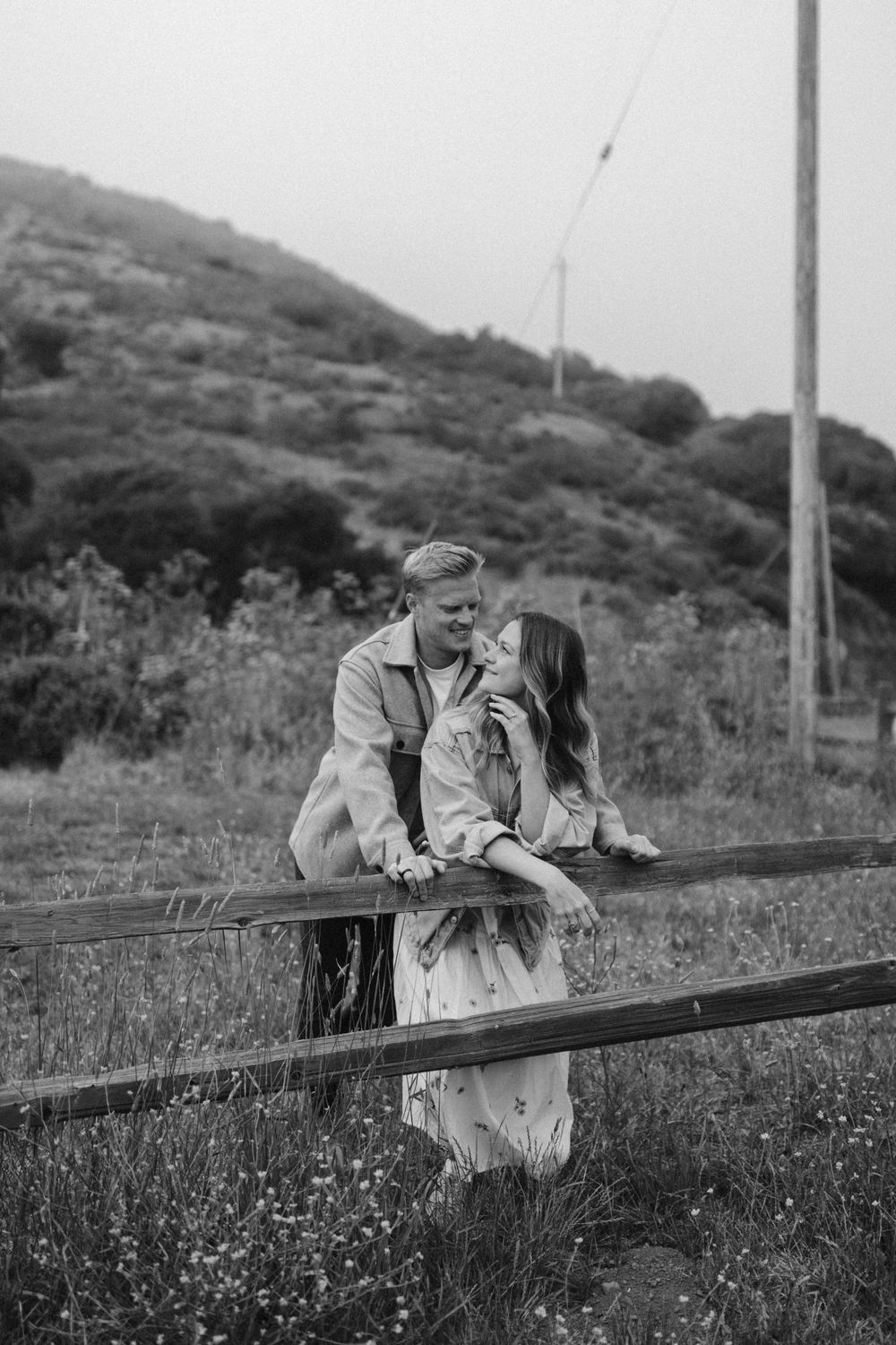 A couple shares a romantic black and white moment leaning against a wooden fence.