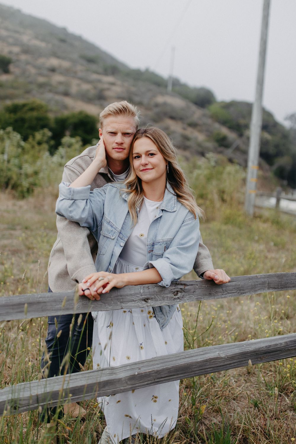A couple poses casually against a wooden fence with rolling hills in the background.