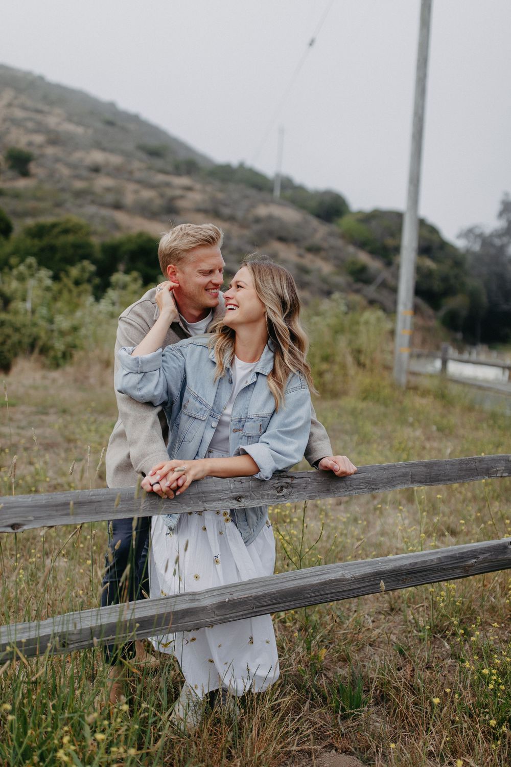 A couple shares an intimate moment while leaning against a wooden fence on a hillside with wild grass.