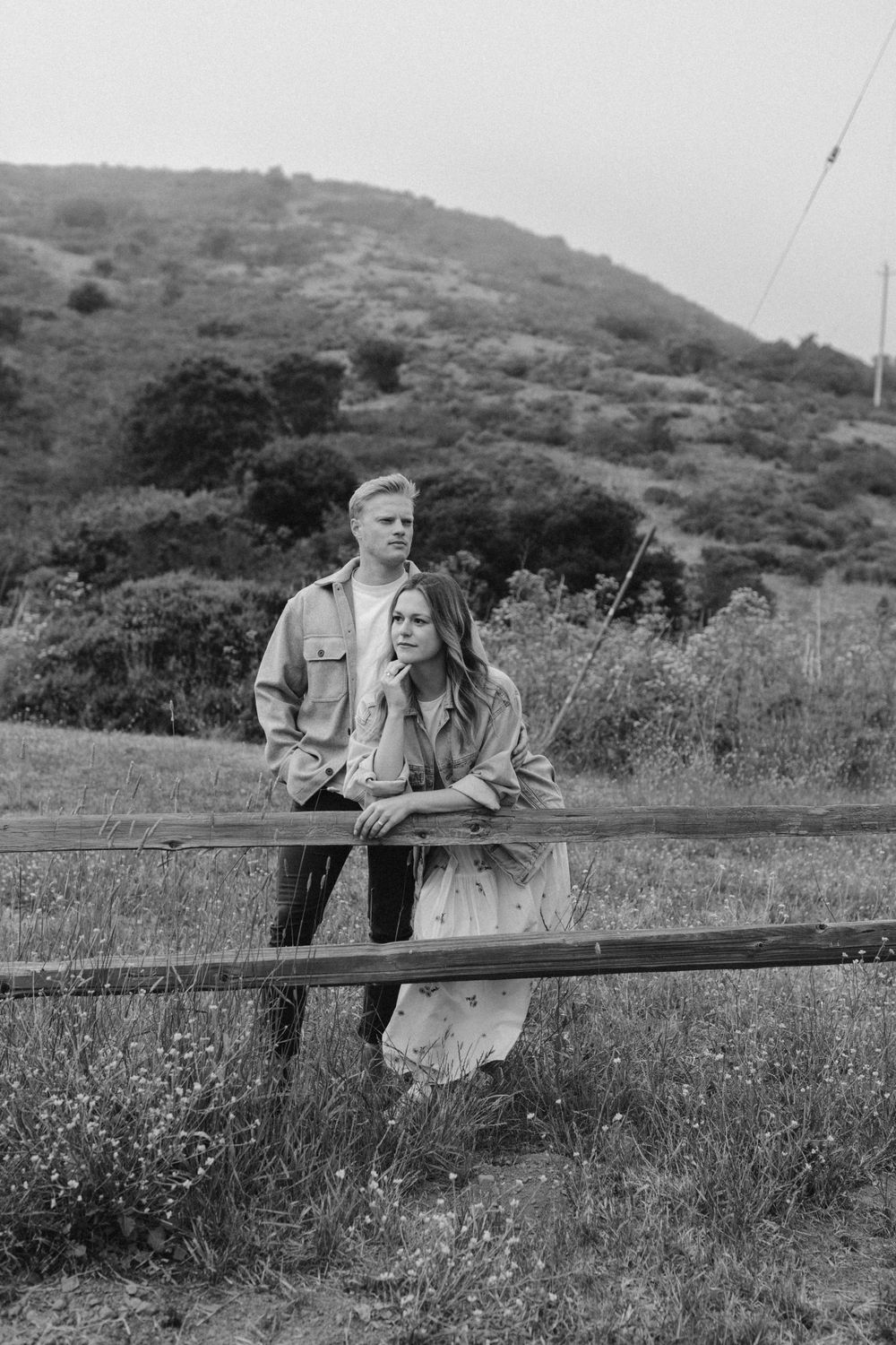 A black and white photo of a couple posing romantically by a rustic fence in a natural landscape setting.