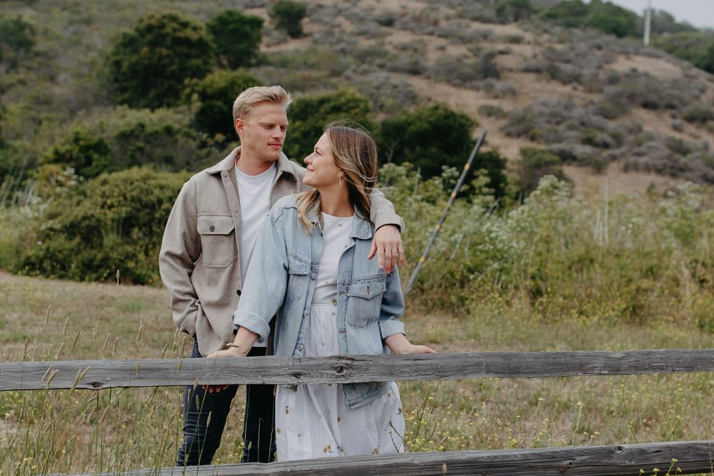 A couple wearing denim jackets embraces by a wooden fence with rolling hills in the background.