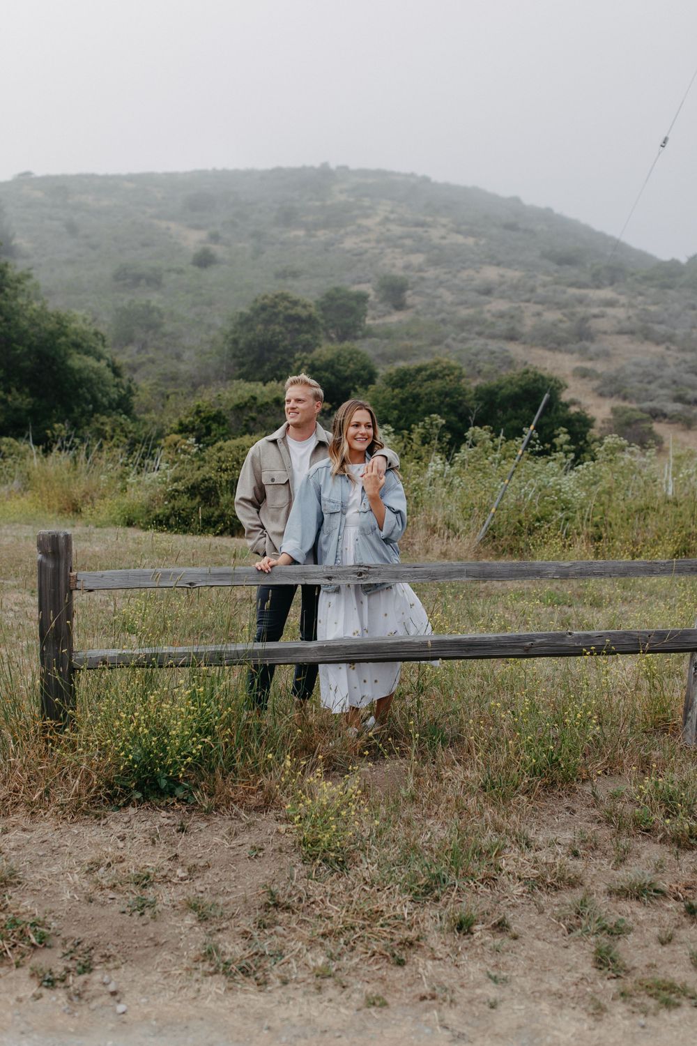 A couple stands together at a wooden fence overlooking a misty hillside landscape.