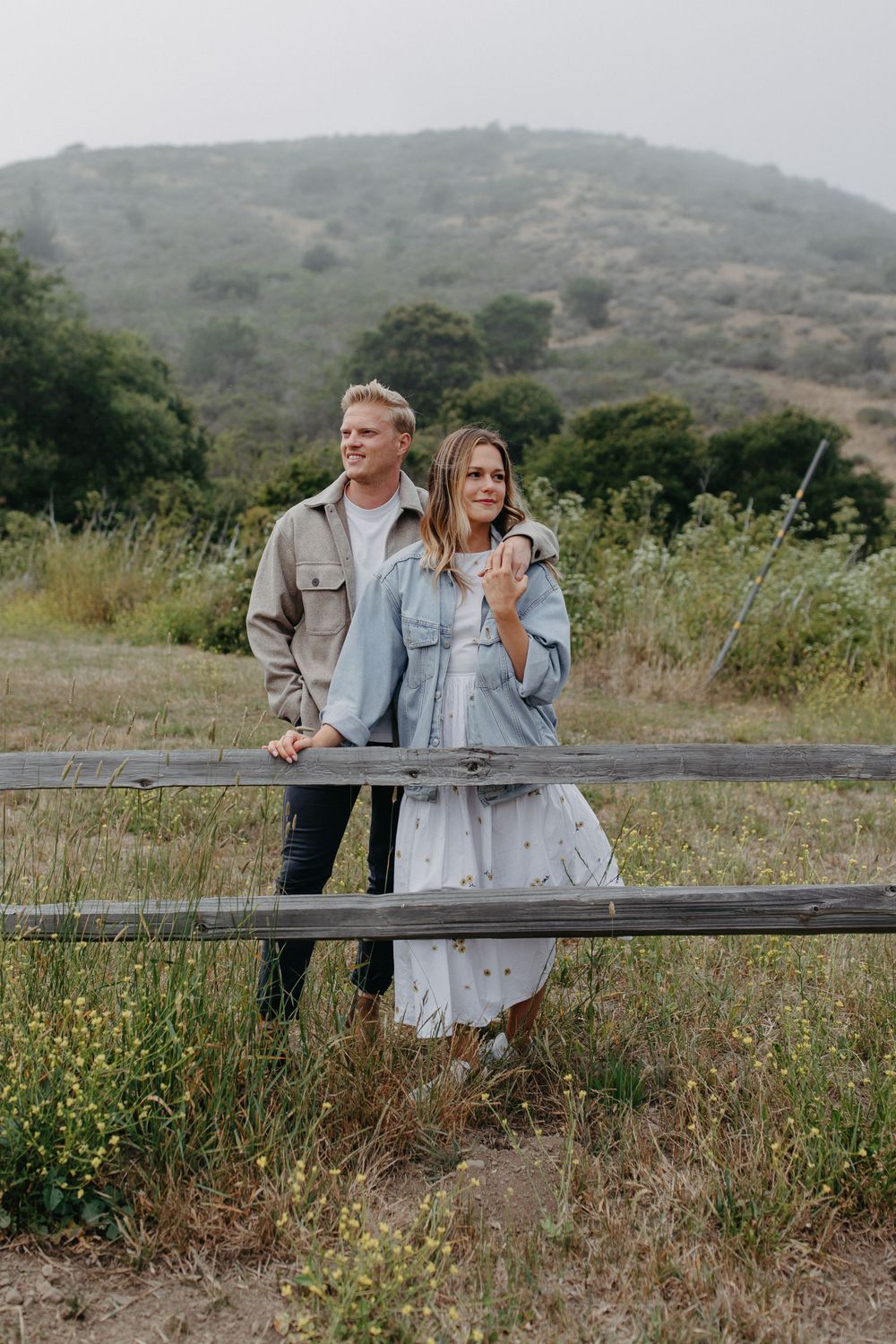 A couple shares a joyful moment while standing at a rustic fence with hills in the background.