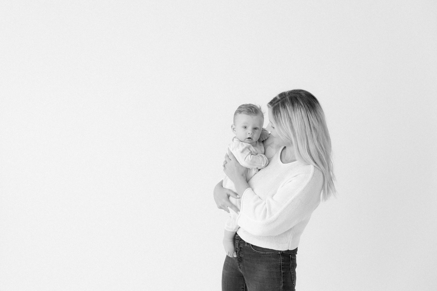 Three month old baby in a white romper is held in mom's arms. Mom looks at baby and baby looks at camera.