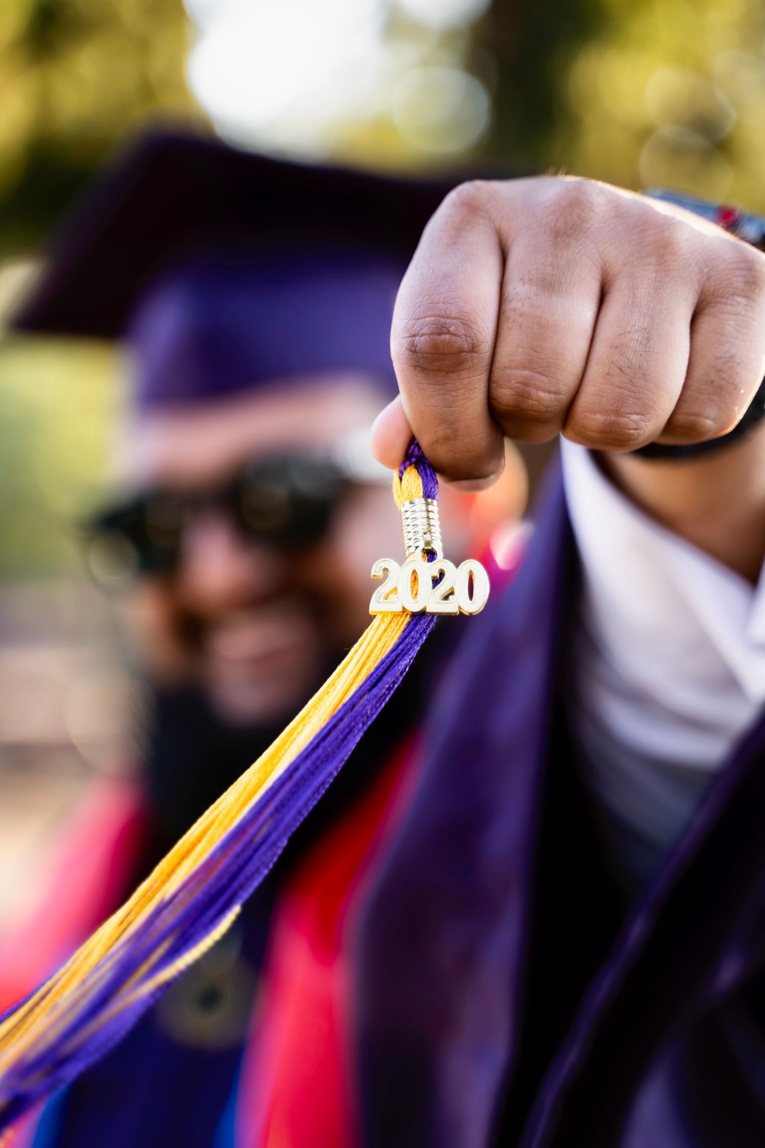 A graduation tassel with '2020' charm dangles against a blurred background with purple and gold ribbons.