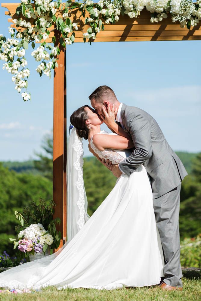 Romantic wedding ceremony kiss under a flower-adorned pergola with mountain views.