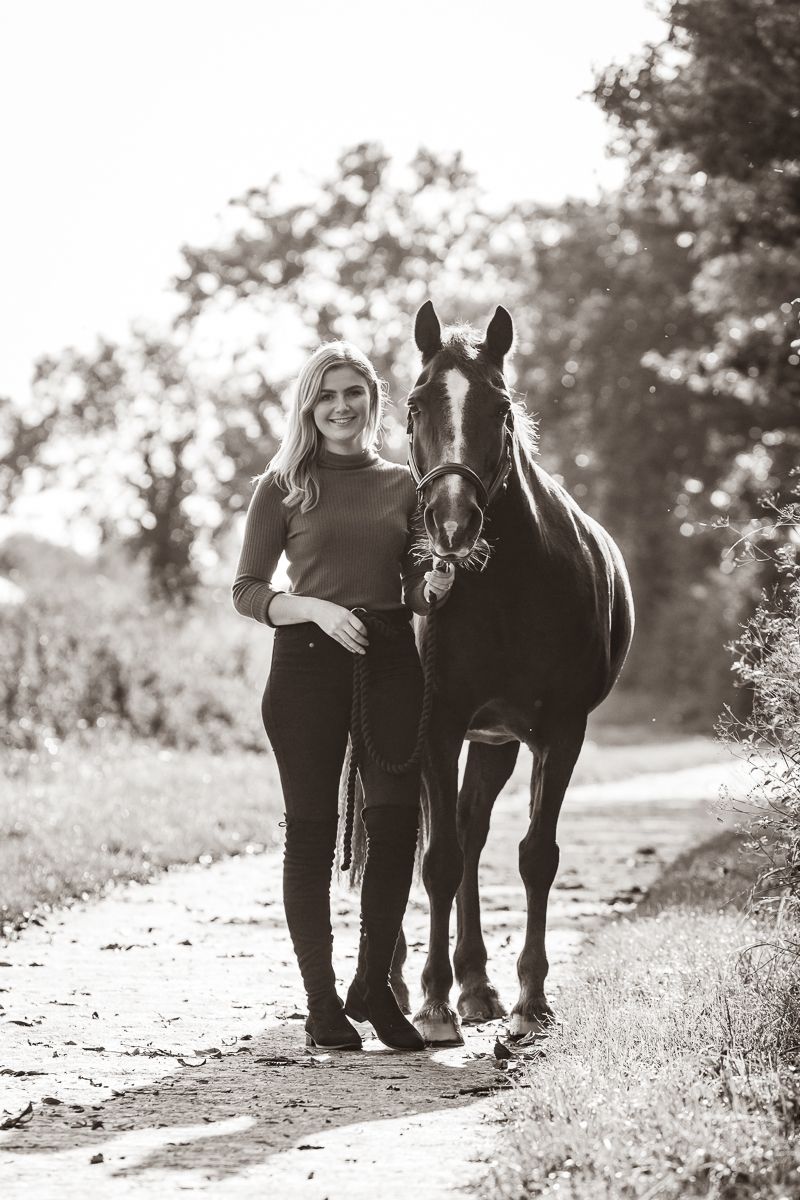 A black and white portrait of a person standing with their dark horse on a country path.