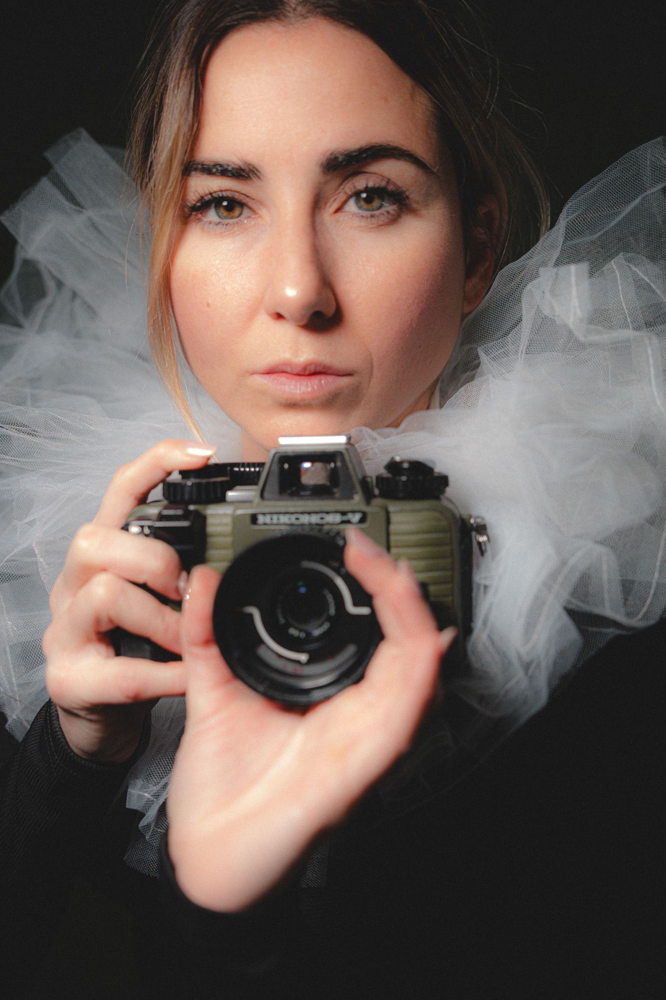 Fine art portrait with vintage camera and tulle dress against dark background.