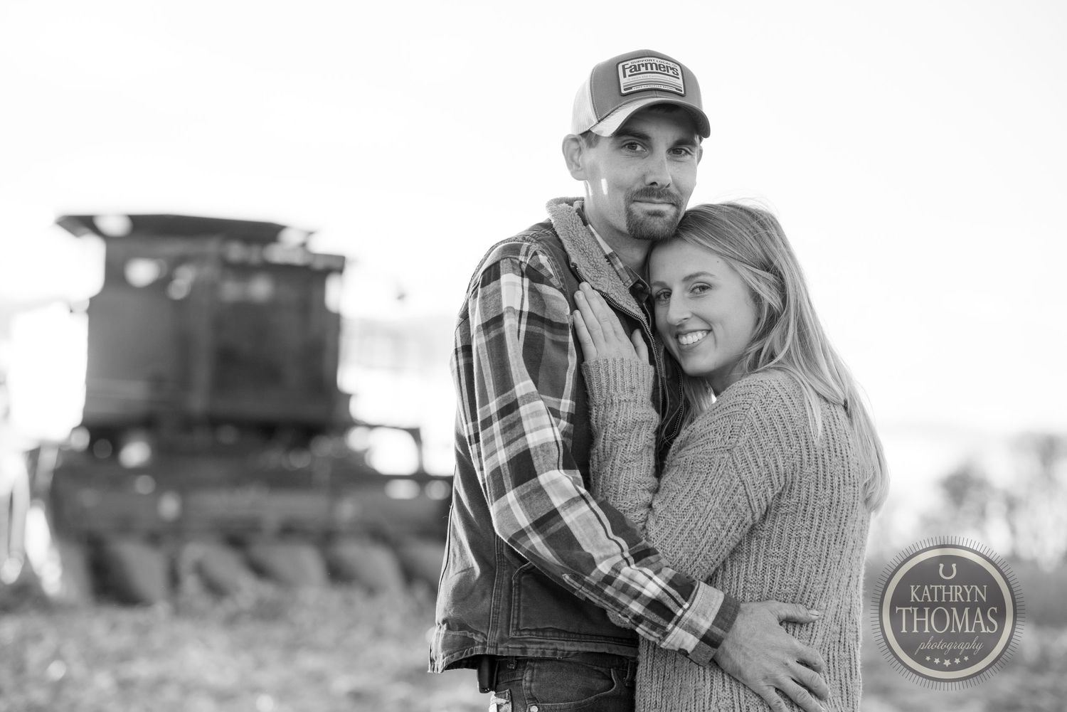 Black and white portrait of an affectionate couple posing with farm equipment in background.