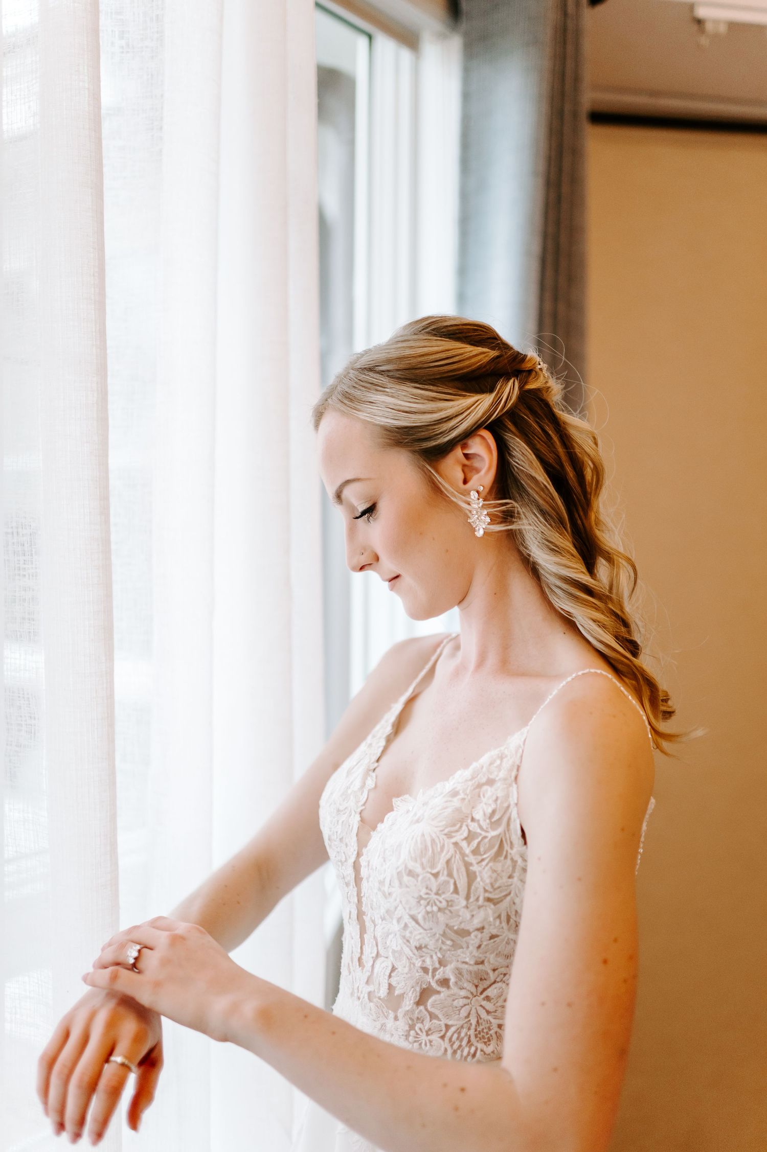 Close-up view of bride in white lace dress preparing for wedding day near bright window.