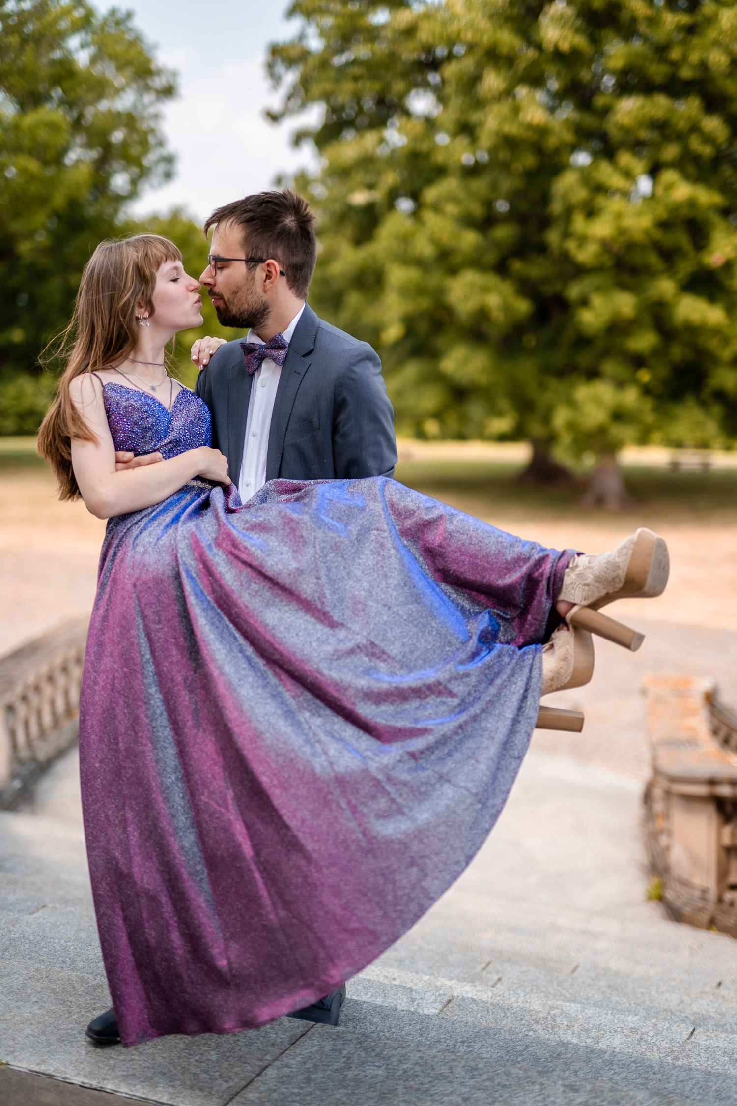 A couple in formal attire embraces on stone steps, with her wearing a flowing purple gown.