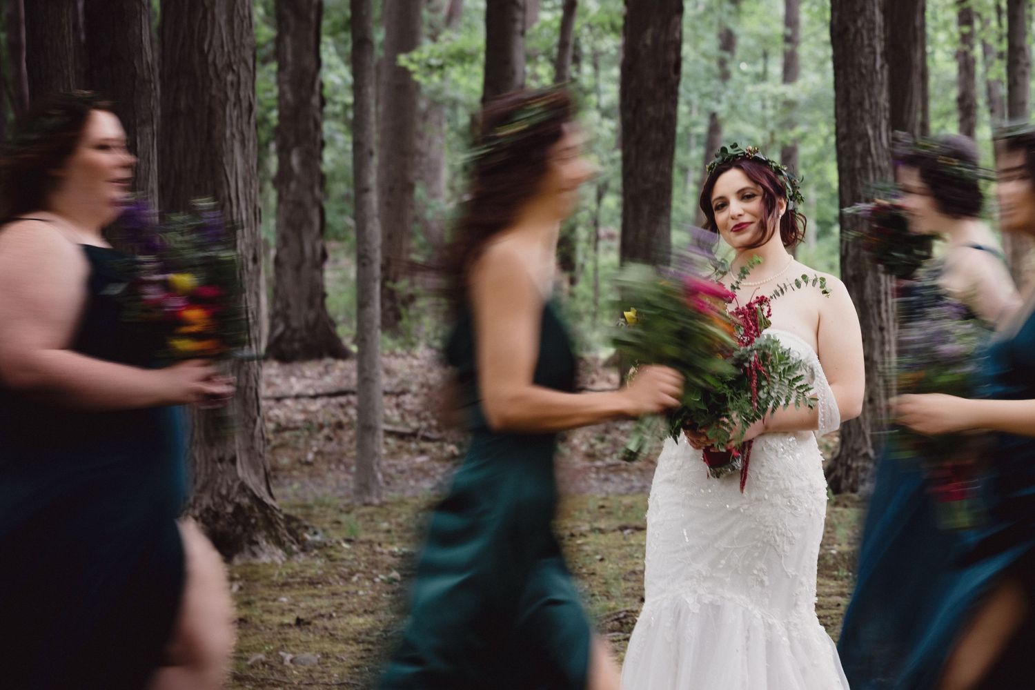 Motion-blurred bridesmaids in teal dresses walk past the bride in a forest setting.
