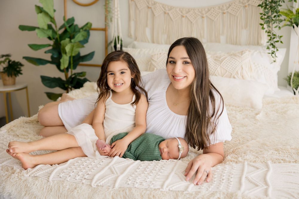 Family portrait on white bed with macrame wall hanging and green plants creating cozy atmosphere.