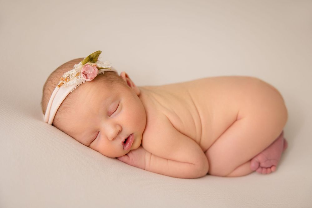 Sleeping newborn baby with floral headband posed in nude position on beige backdrop.