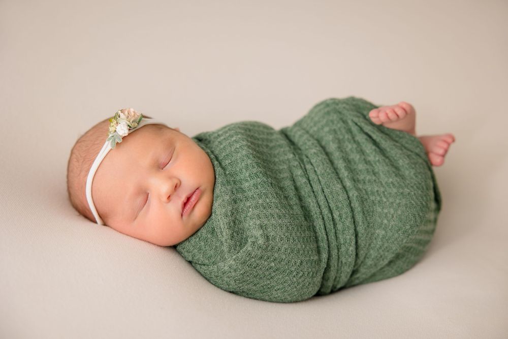 Peaceful sleeping newborn wrapped in sage green knit wrap wearing delicate headband.