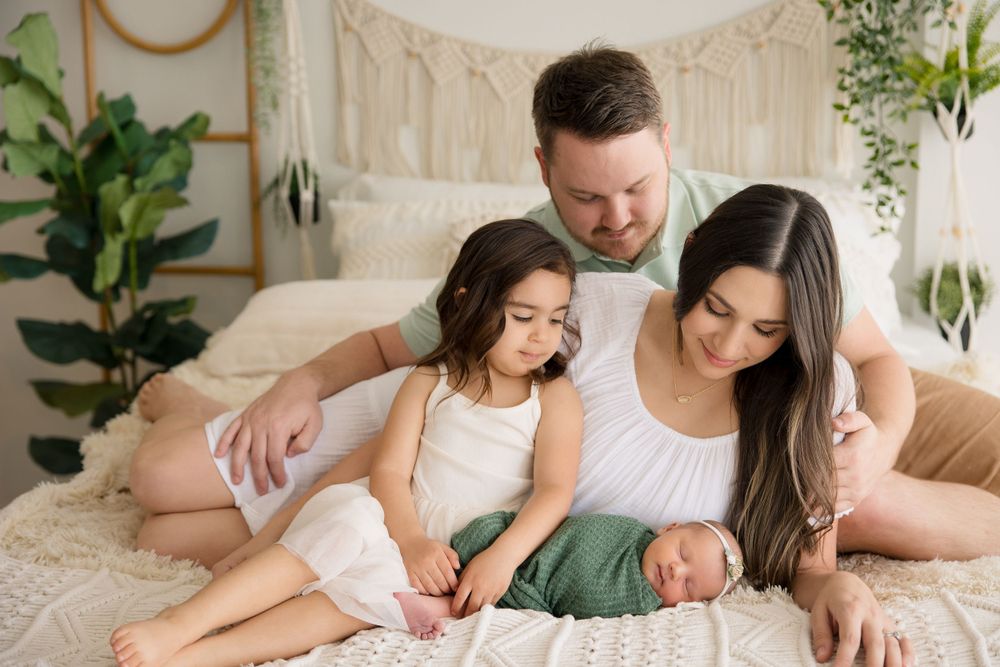 Intimate family moment captured on white bedding with macrame wall hanging and indoor plants.