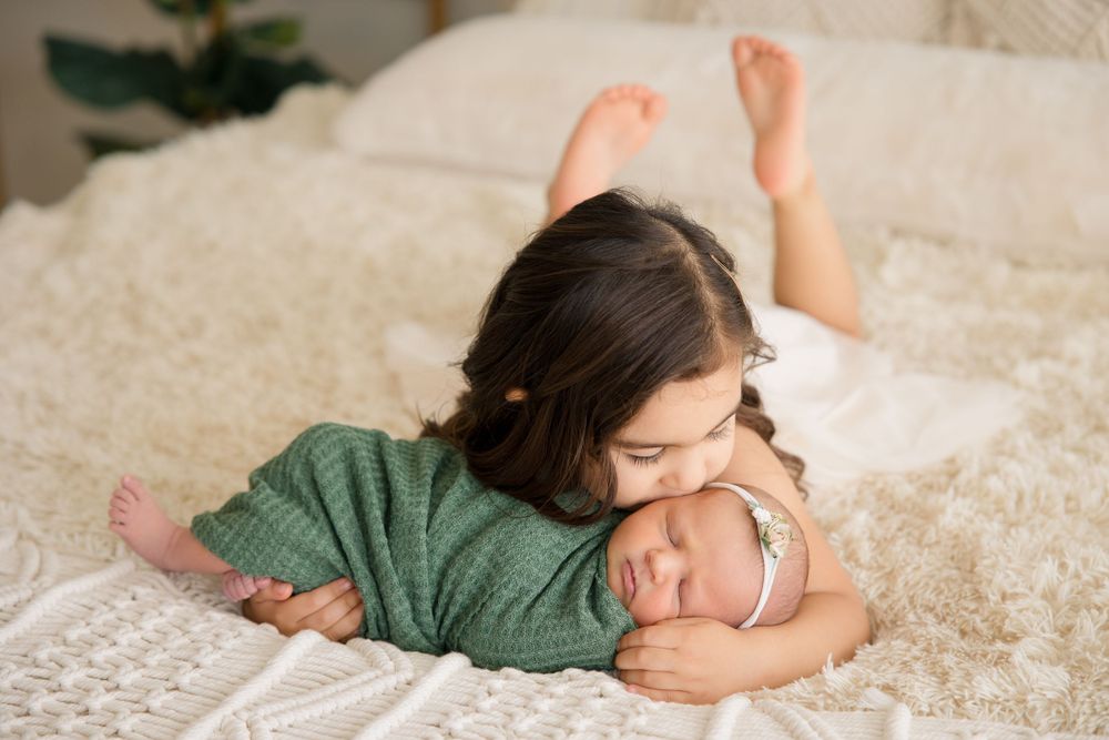 Two siblings share a tender moment lying together on a white bed with macrame wall decor and plants in background.