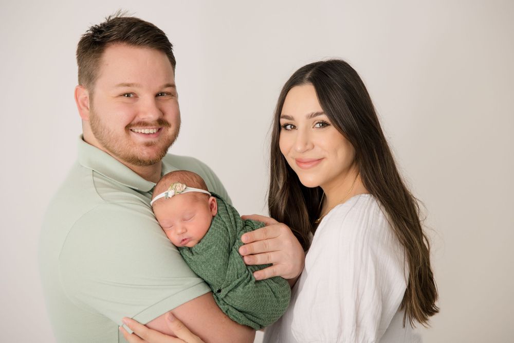 A family portrait with a newborn baby wrapped in green between two parents on a light background.