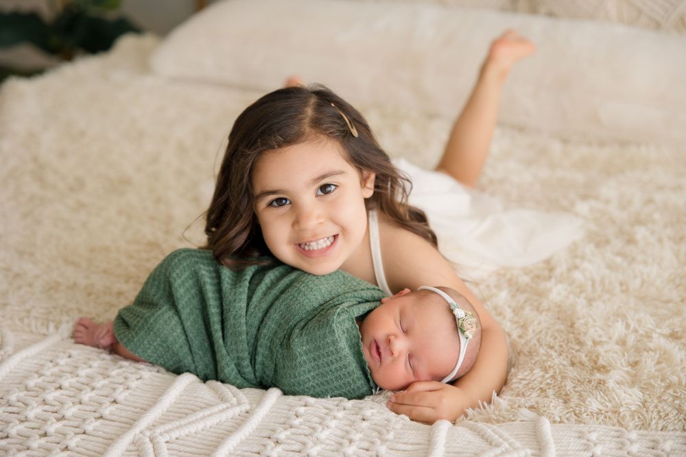 Sibling bonding moment captured on cozy white bed with decorative wall hanging in background.
