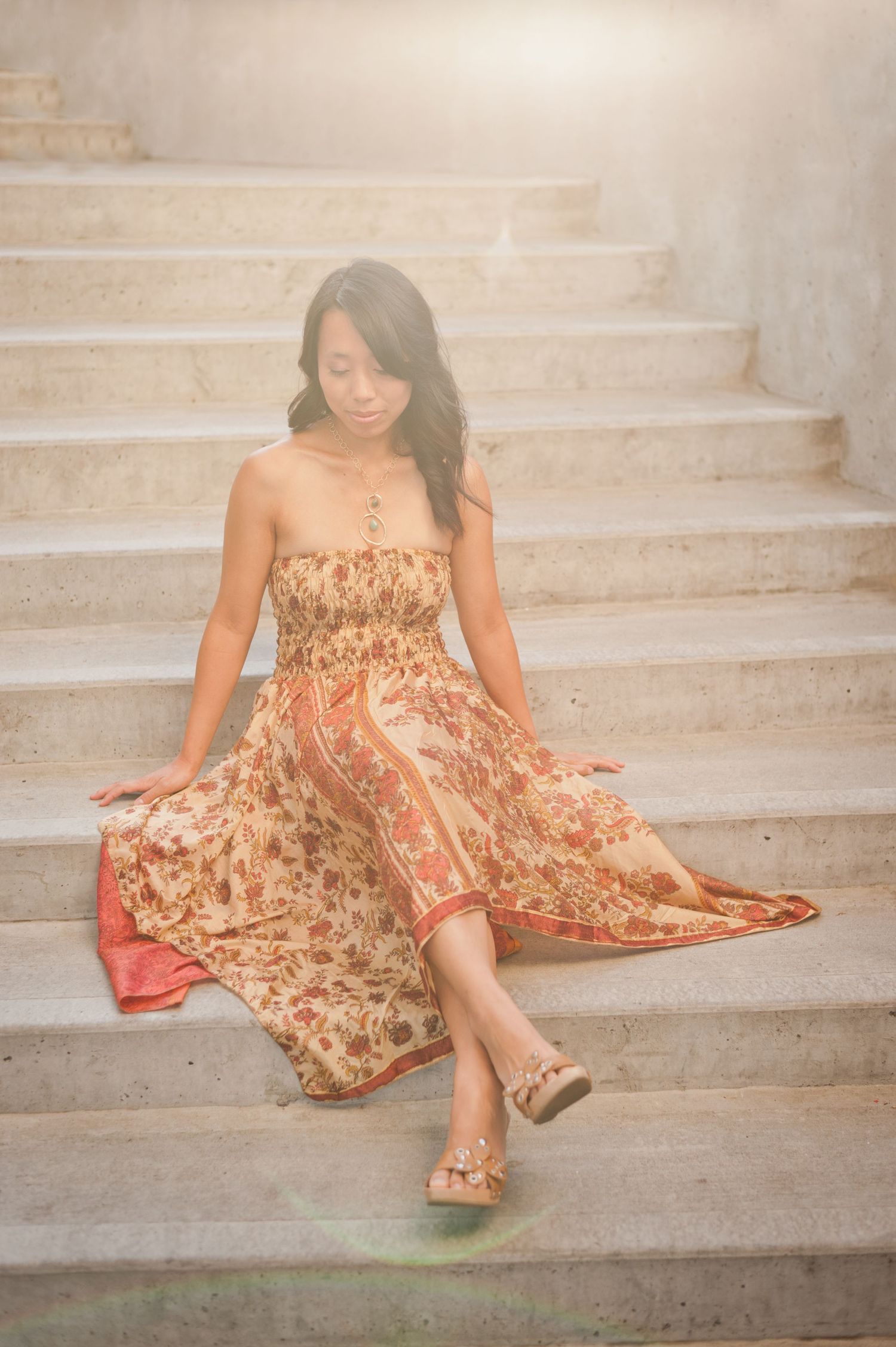 A woman in a flowing floral dress sits gracefully on concrete steps in soft sunlight.