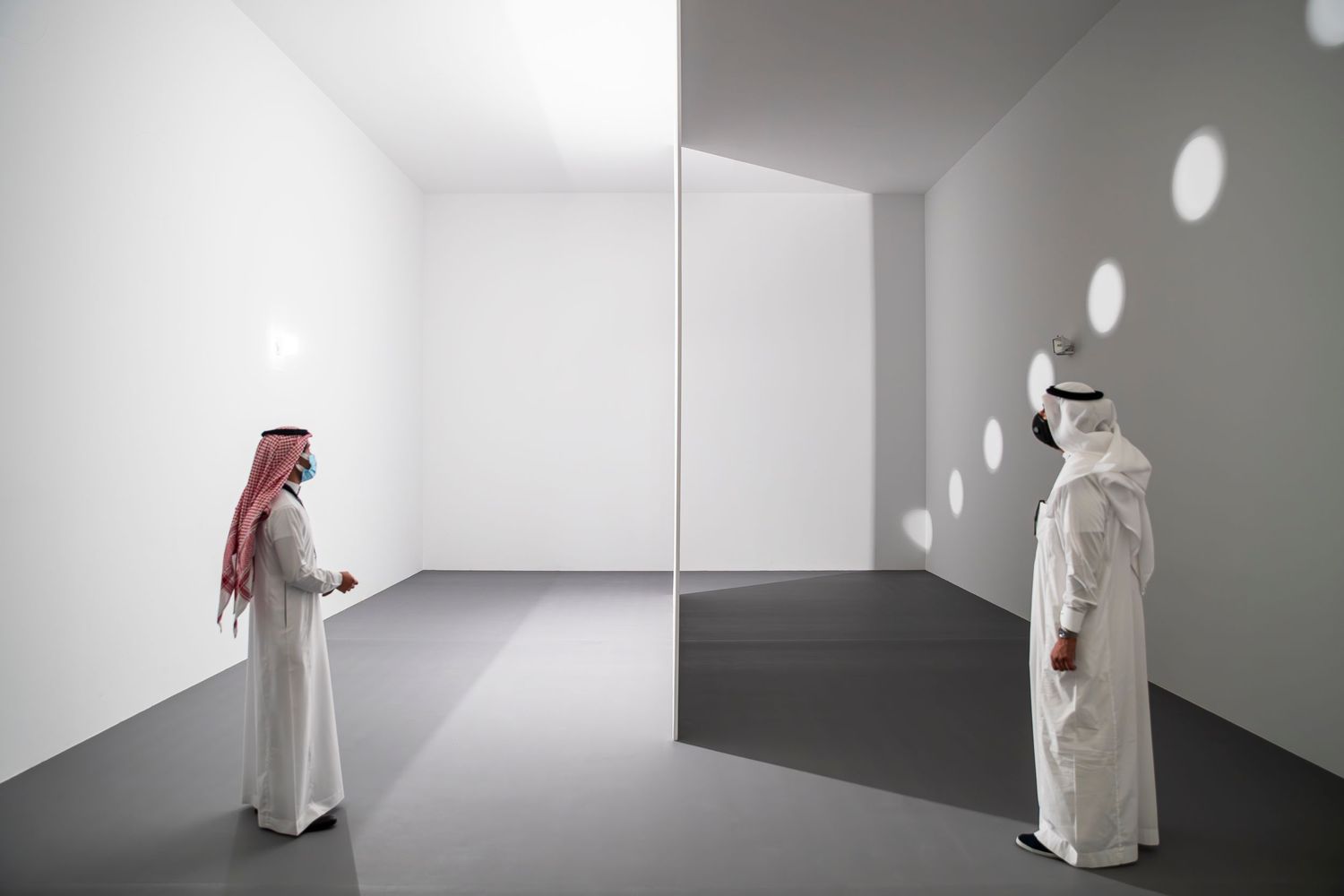 Two people in traditional Gulf dress examine a minimalist white gallery installation.