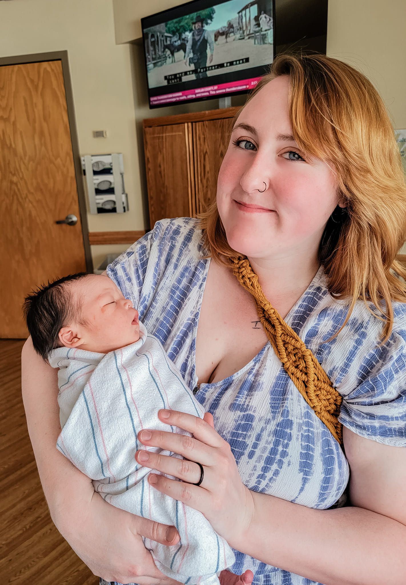 New mother with nose ring smiling while holding swaddled newborn baby indoors.