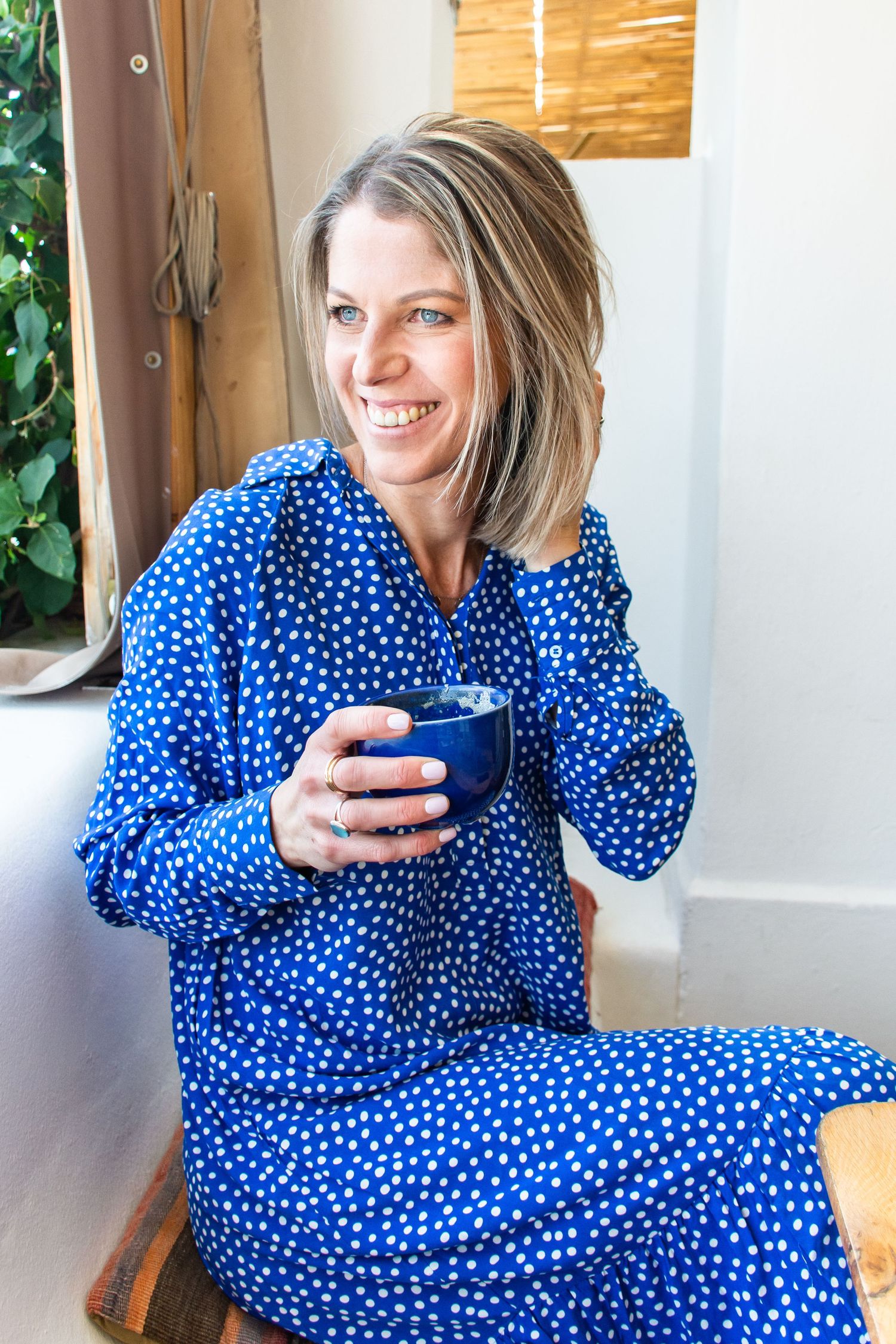 Person relaxing in blue polka dot dress while holding coffee cup by window.