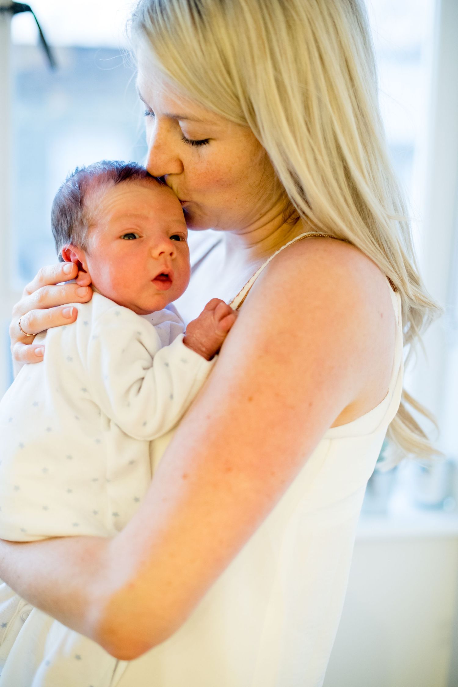 Woman in white tank top gently kisses infant wearing cream colored outfit in tender maternal moment.